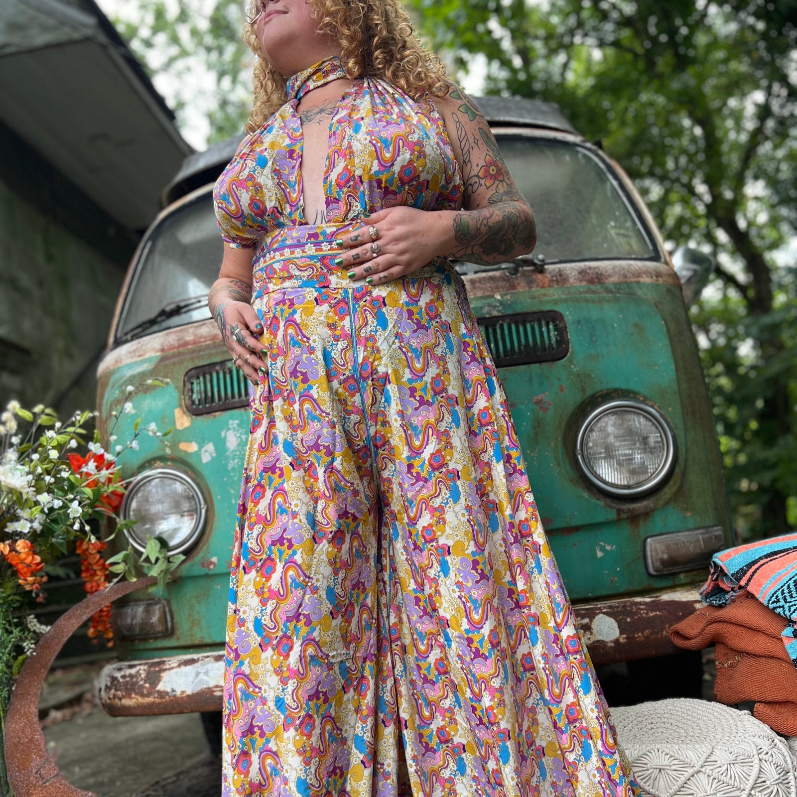 Person wearing a colorful floral one piece outfit standing in front of a vintage van with greenery around.