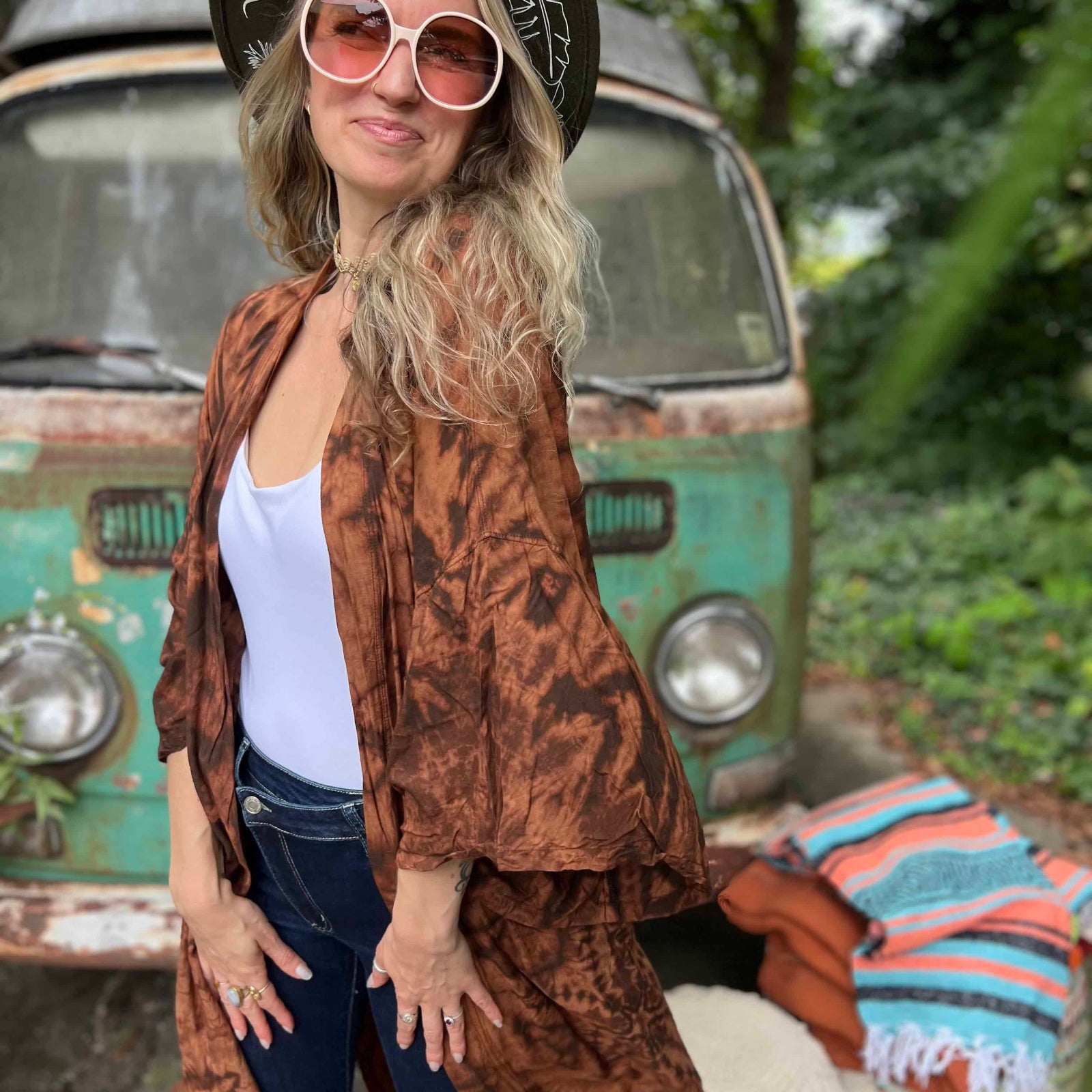 Woman in a patterned jacket and hat standing in front of a vintage van with a colorful blanket.