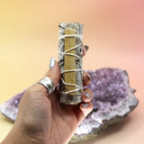 The White Sage with Palo Santo Smudge Stick 4 in. being held in front of an amethyst piece on a pink and yellow backround.