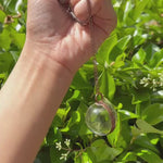 Someone holding the Crystal Ball Necklace in Copper - Clear Quartz in front of greenery.