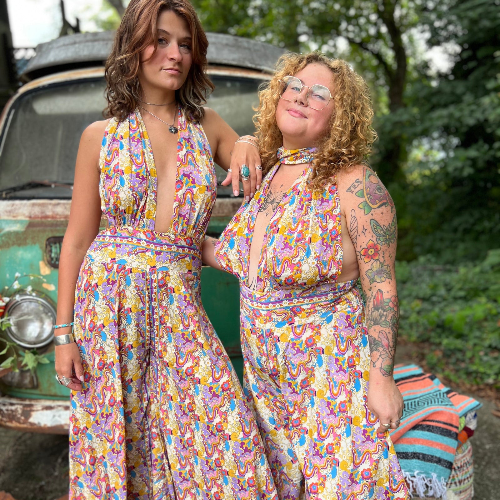 Two women in colorful floral jumpsuits standing next to a vintage car with a green leafy background.