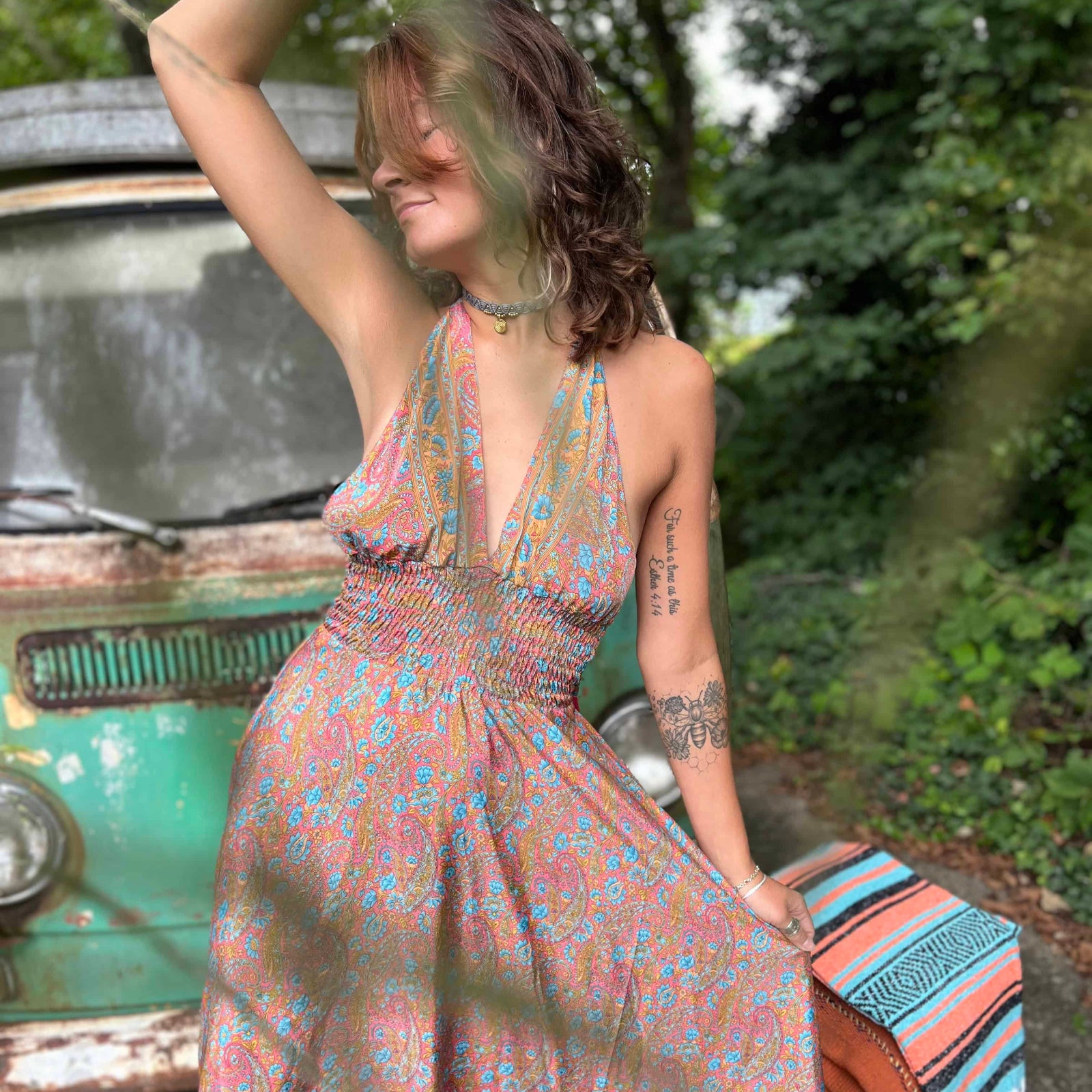 Woman in a floral dress sitting next to a vintage car with greenery in the background