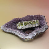 The White Sage & Sweetgrass Smudge Stick 4 in. on an amethyst piece on a pink and yellow background.