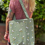 Blonde woman wearing a floral dress and carrying a sage green linen tote bag with hand-embroidered white daisies