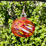A colorful handblown art glass hummingbird feeder hanging from a metal loop, with a blurred greenery background.