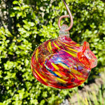 A colorful handblown art glass hummingbird feeder hanging from a metal loop, with a blurred greenery background.