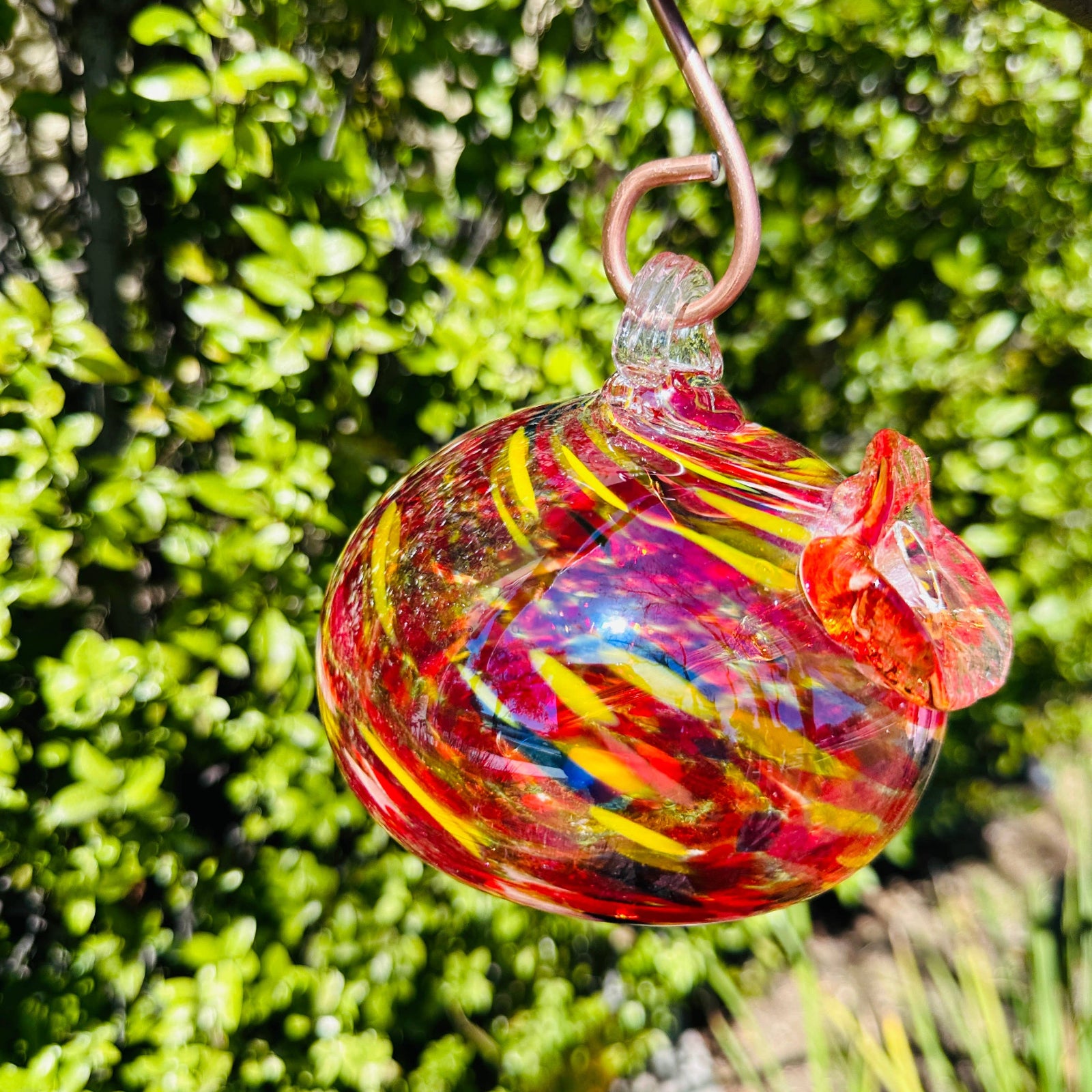 A colorful handblown art glass hummingbird feeder hanging from a metal loop, with a blurred greenery background.