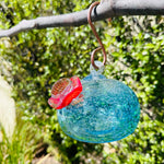 A colorful handblown art glass hummingbird feeder hanging from a metal loop, with a blurred greenery background.