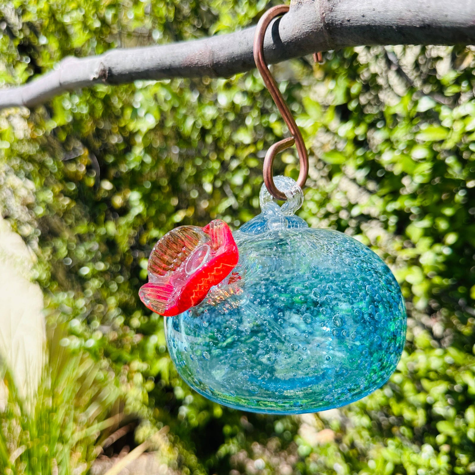 A colorful handblown art glass hummingbird feeder hanging from a metal loop, with a blurred greenery background.