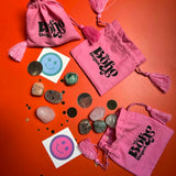 Pink drawstring bags with 'The Boho Den' branding, stones, and smiley face stickers on an orange background.