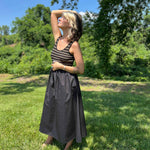 Full-body view of woman in striped knit and black midi dress with one arm raised, enjoying the sunshine.