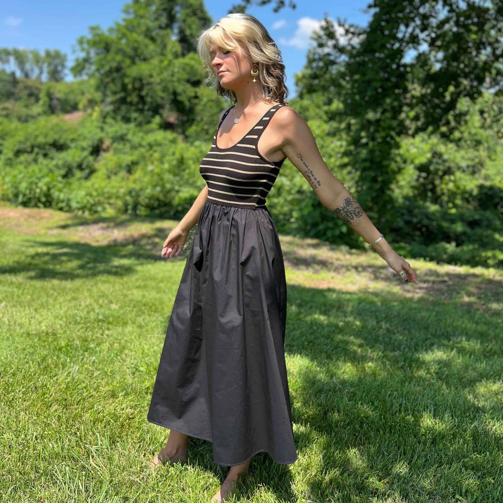 Side profile of woman in a striped tank dress with A-line black skirt, standing barefoot on grass.