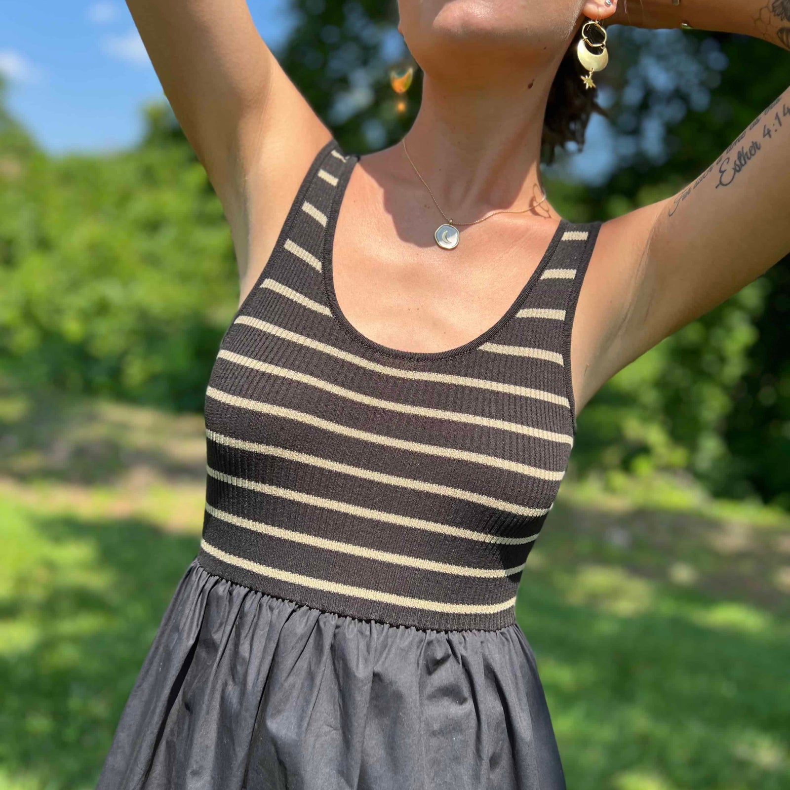 Woman with tattoos standing barefoot in a field wearing a striped ribbed top and long black skirt dress.