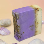 Purple soap bar with natural texture surrounded by small stones and crystals on a peach background