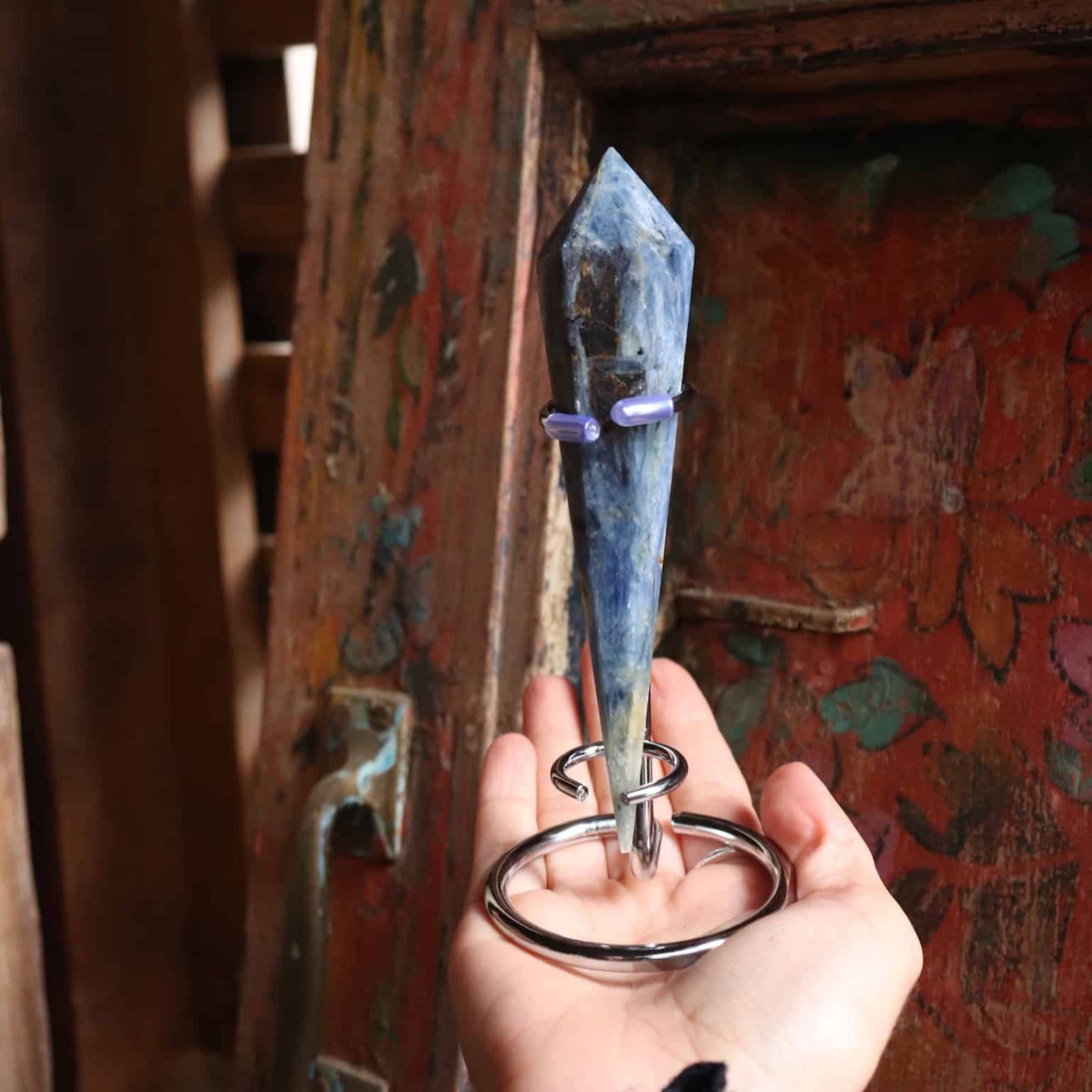 Hand holding a blue kyanite crystal on a stand in front of a rustic wooden door with peeling paint.