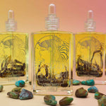 3 bottles standing up of essence of hathor blue lotus oil on a pink and orange pastel background with turquoise stones scattered in front