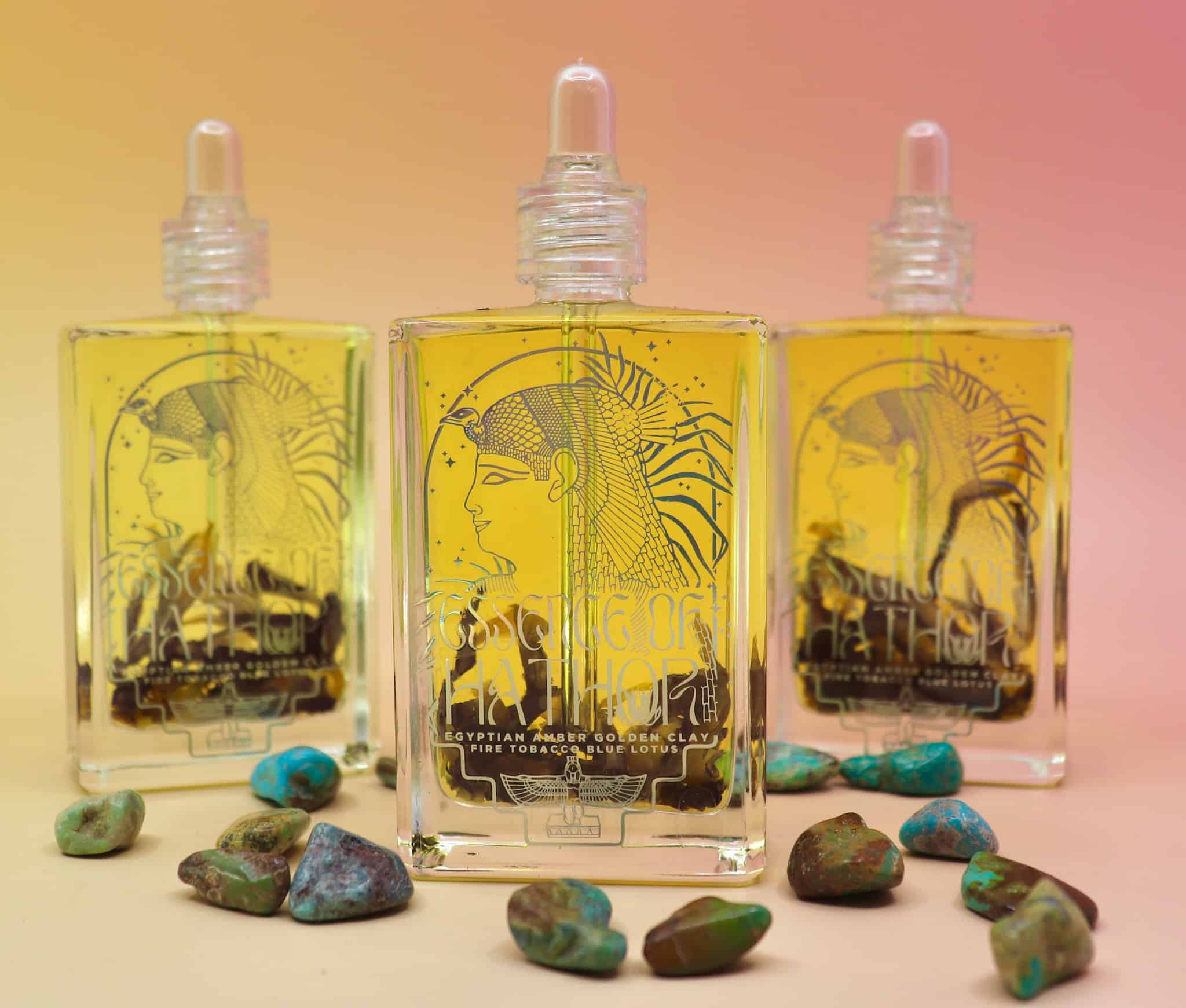 3 bottles standing up of essence of hathor blue lotus oil on a pink and orange pastel background with turquoise stones scattered in front