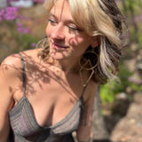 close up of model wearing beautiful gold circle and triangle earrings and wearing the striped blue sundress with pink flowers and trees in background