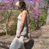 side view of model wearing a teal striped sundress with blonde bangs and brown hair in front of pink trees and woods