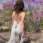 back view of model wearing teal striped sundress with tie in back walking towards pink trees in forest 