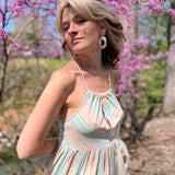 model wearing a halter striped sundress teal color and read with head to the side and slight smile in front of pink trees