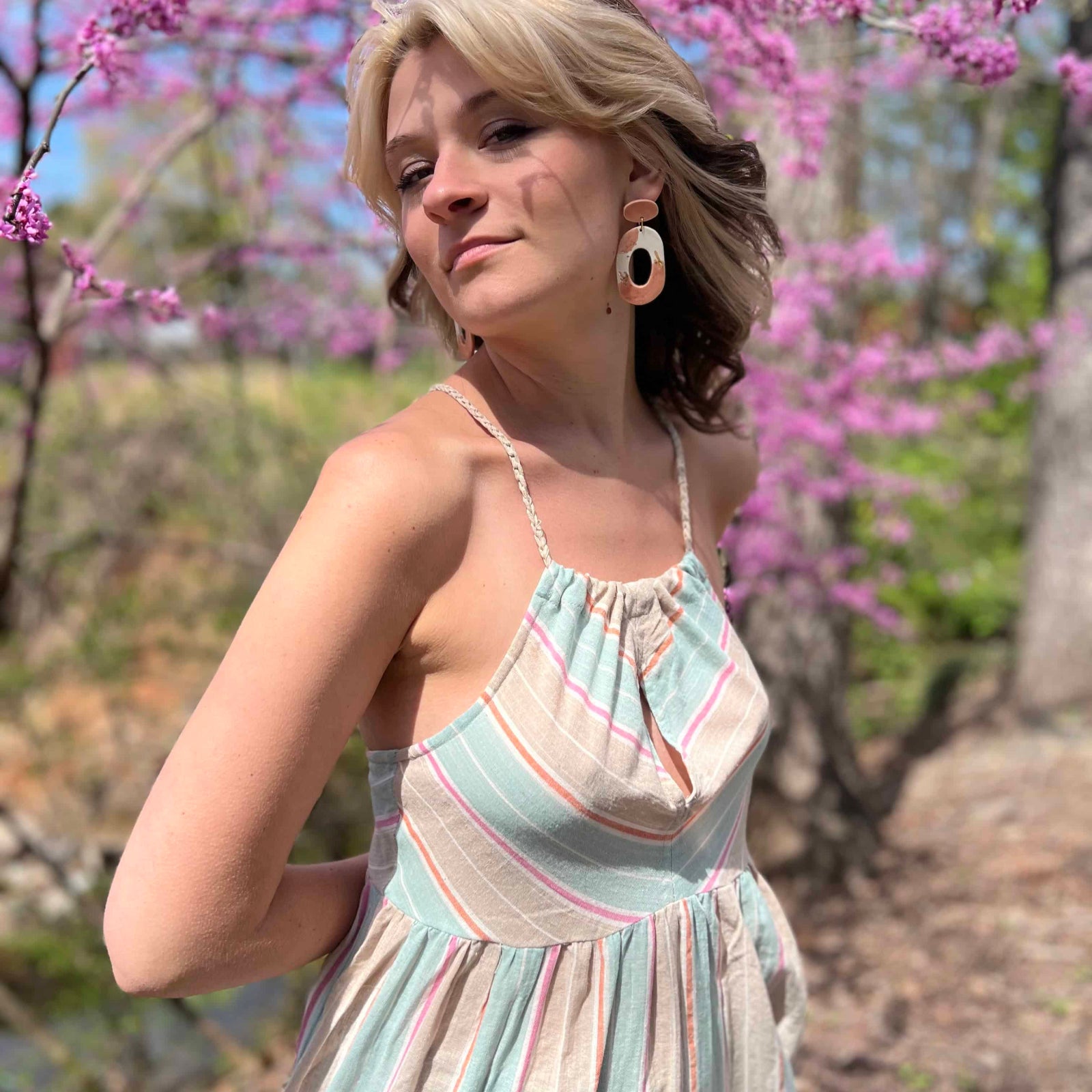model wearing a halter striped sundress teal color and read with head to the side and slight smile in front of pink trees