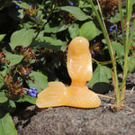 Orange calcite crystal resembling a stylized face on a stone surface with greenery in the background