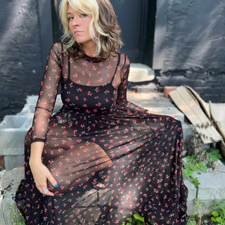 Woman sitting on a cinderblock step in a sheer cherry print maxi dress with crossed arms and soft lighting holding out the side of her dress