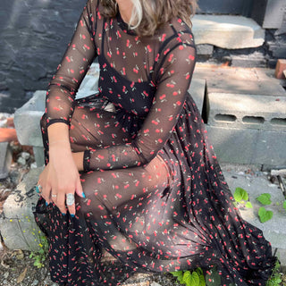 Woman sitting on a cinderblock step in a sheer cherry print maxi dress with crossed arms and soft lighting.