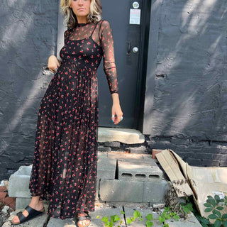Woman in a cherry print mesh maxi dress standing on cement blocks in front of a black door