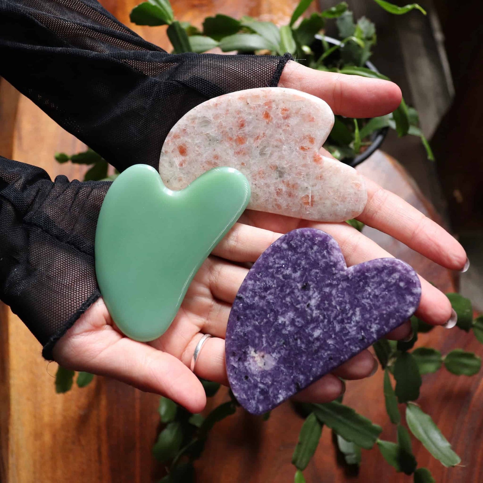 Three heart-shaped stones in green, white, and purple held in a hand with a blurred plant background.