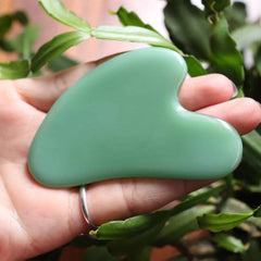 Green aventurine gua sha tool held in a hand with a blurred green plant background