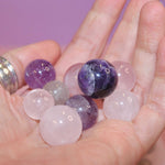 A hand holding various polished crystal marbles, including purple amethyst and clear quartz.