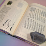 "The Green Witch's Garden" book on a pink and blue background, open and showing some of its pages.