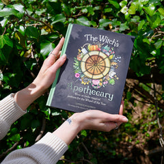 Someone holding the Witch's Apothecary: Seasons of the Witch Book in front of green leaves.