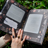 Someone holding the Witch's Apothecary: Seasons of the Witch Book in front of green leaves. The book is open showing some pages.