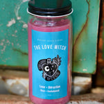 The Love Witch Spell Candle with Herbs and Crystals in front of a teal background and on a white rusty surface.