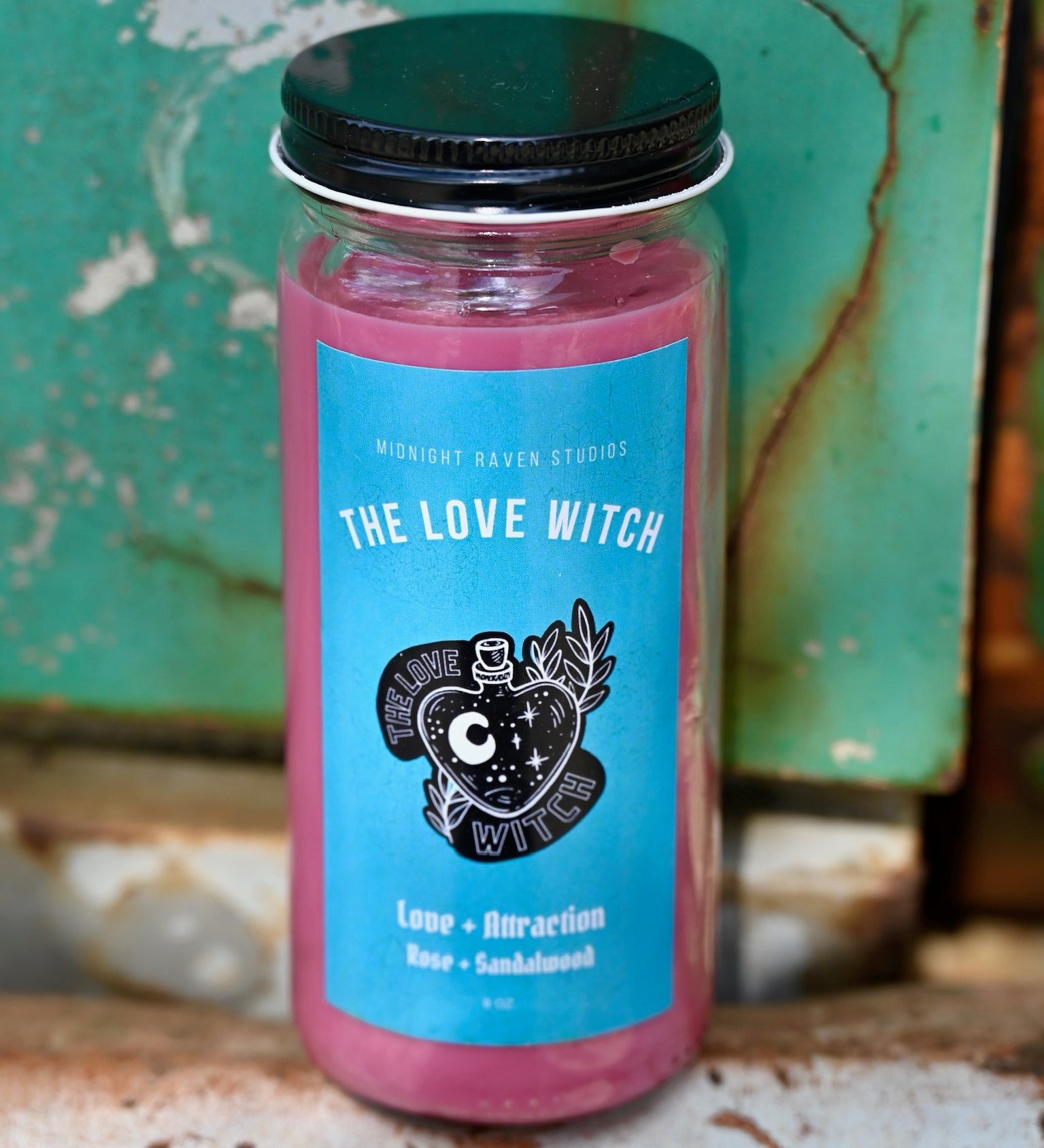 The Love Witch Spell Candle with Herbs and Crystals in front of a teal background and on a white rusty surface.