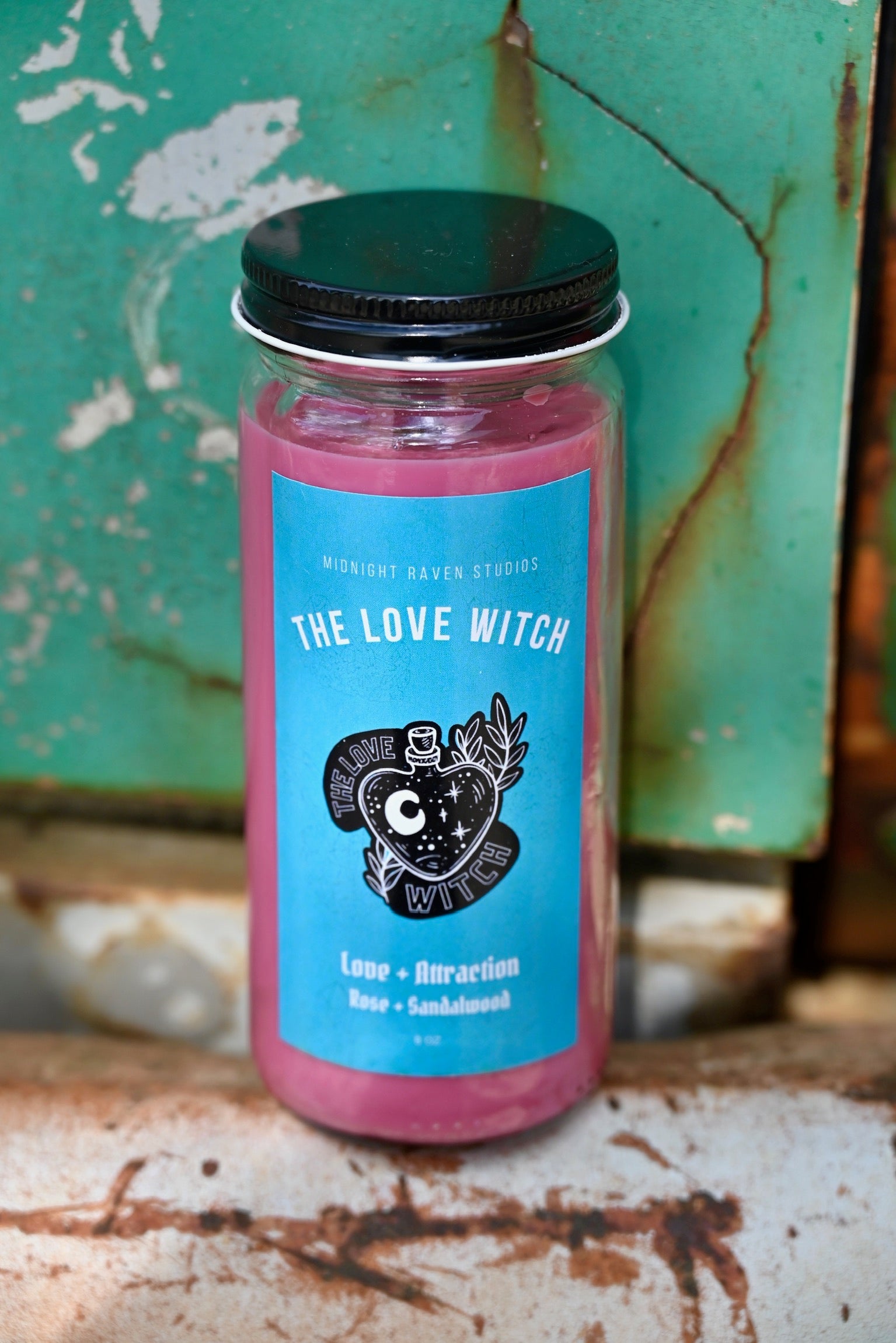 The Love Witch Spell Candle with Herbs and Crystals in front of a teal background and on a white rusty surface.