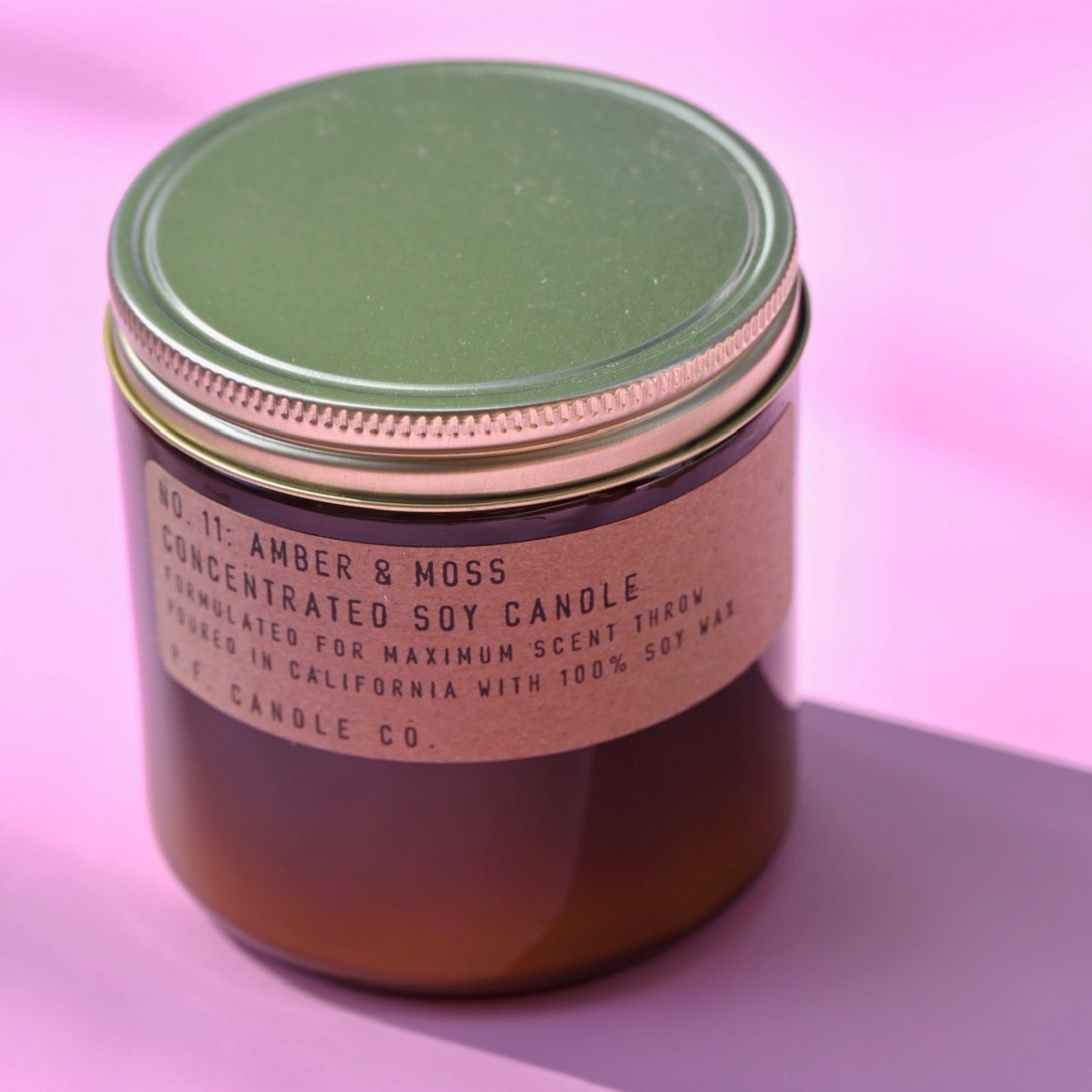 The P.F. Candle Co. - Amber & Moss - 12.5 oz Concentrated Soy Candle on a pink background.
