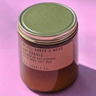 The P.F. Candle Co. - Amber & Moss - 7.2 oz Standard Soy Candle on a pink background.