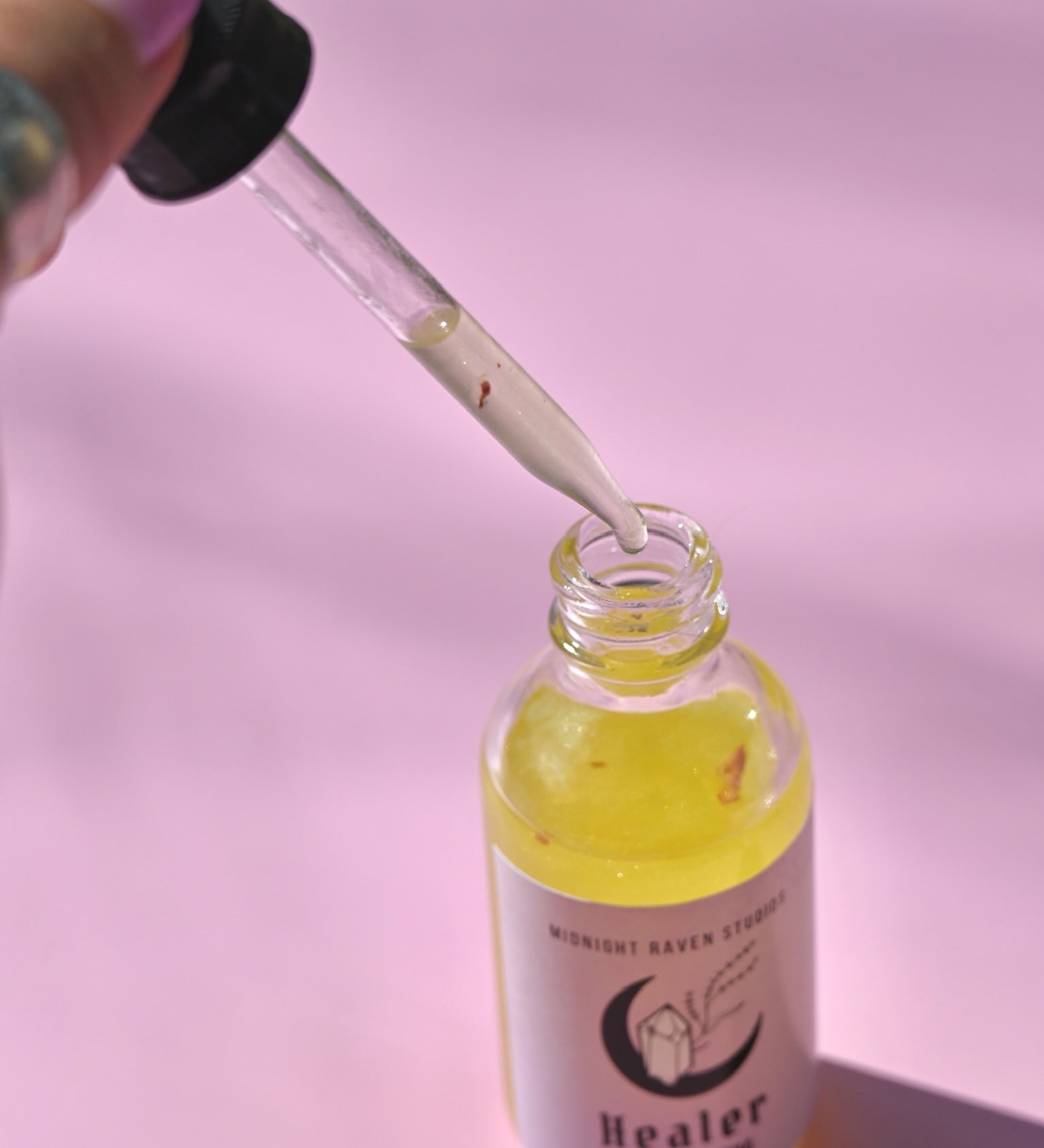 The Healer Potion Body Oil - Lavender Rose Body Oil on a pink background with someone holding the dropper next to the bottle.