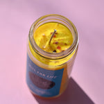 The Zest for Life Spell Candle with Herbs and Crystals on a pink background with the lid off showing the inside of the candle.