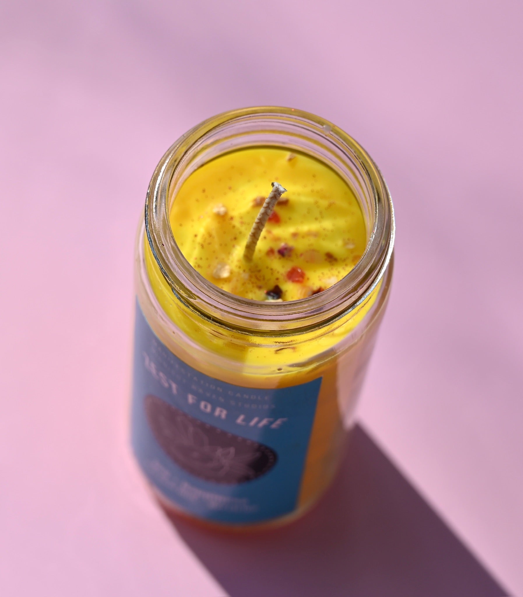 The Zest for Life Spell Candle with Herbs and Crystals on a pink background with the lid off showing the inside of the candle.