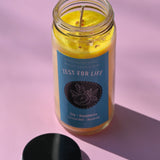 The Zest for Life Spell Candle with Herbs and Crystals on a pink background with its lid open and to the side.
