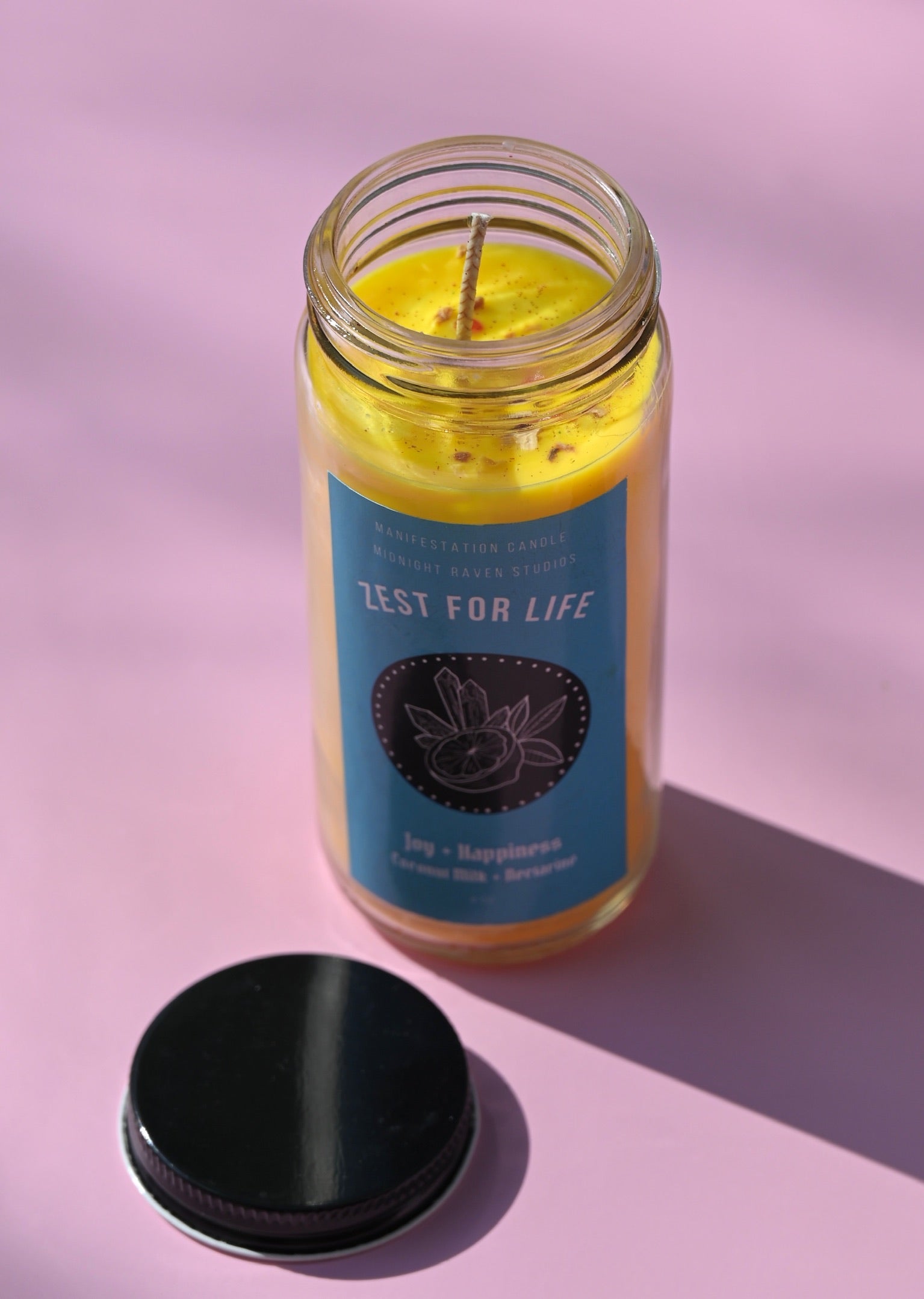 The Zest for Life Spell Candle with Herbs and Crystals on a pink background with its lid open and to the side.