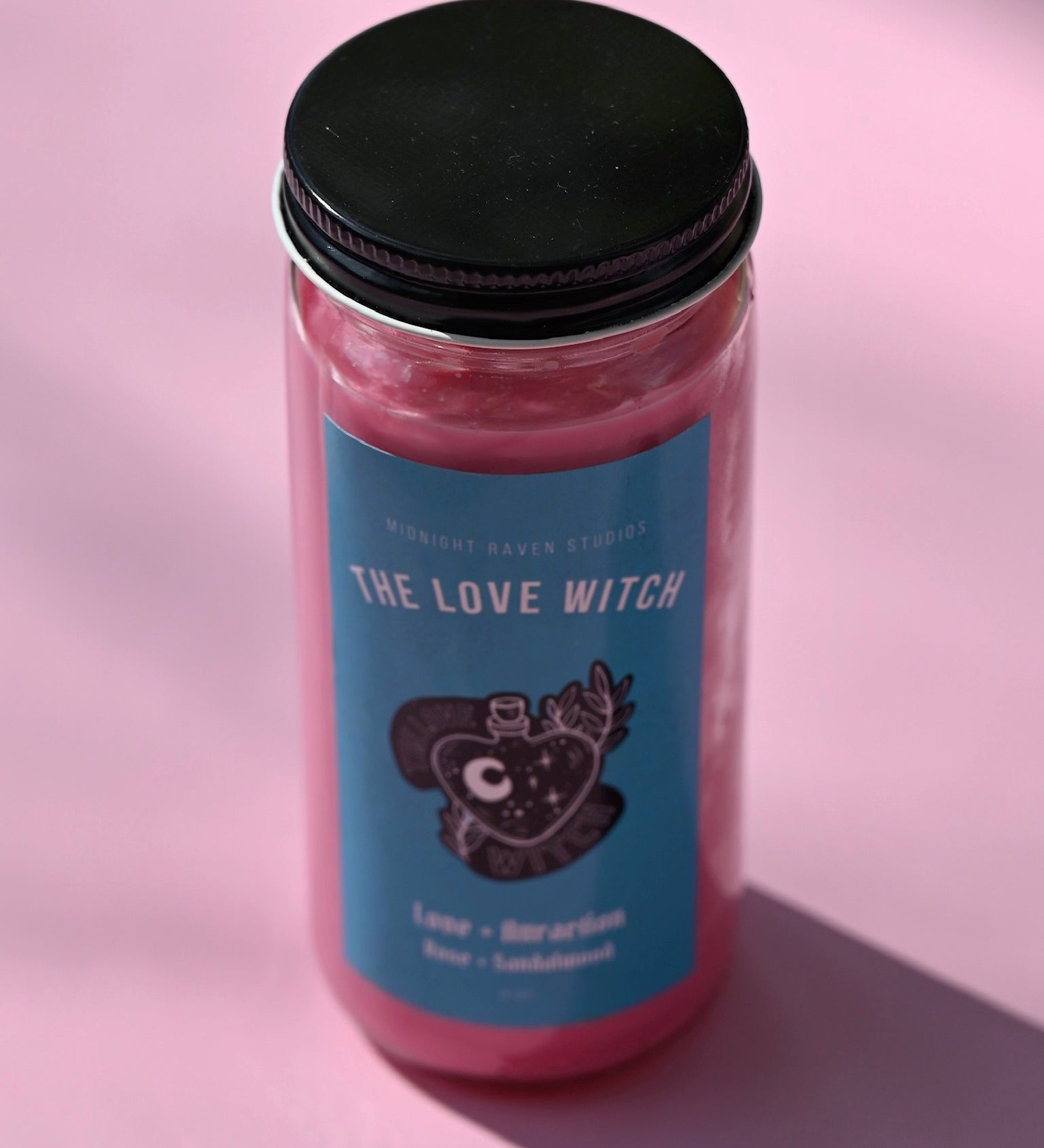 The Love Witch Spell Candle with Herbs and Crystals on a pink background.