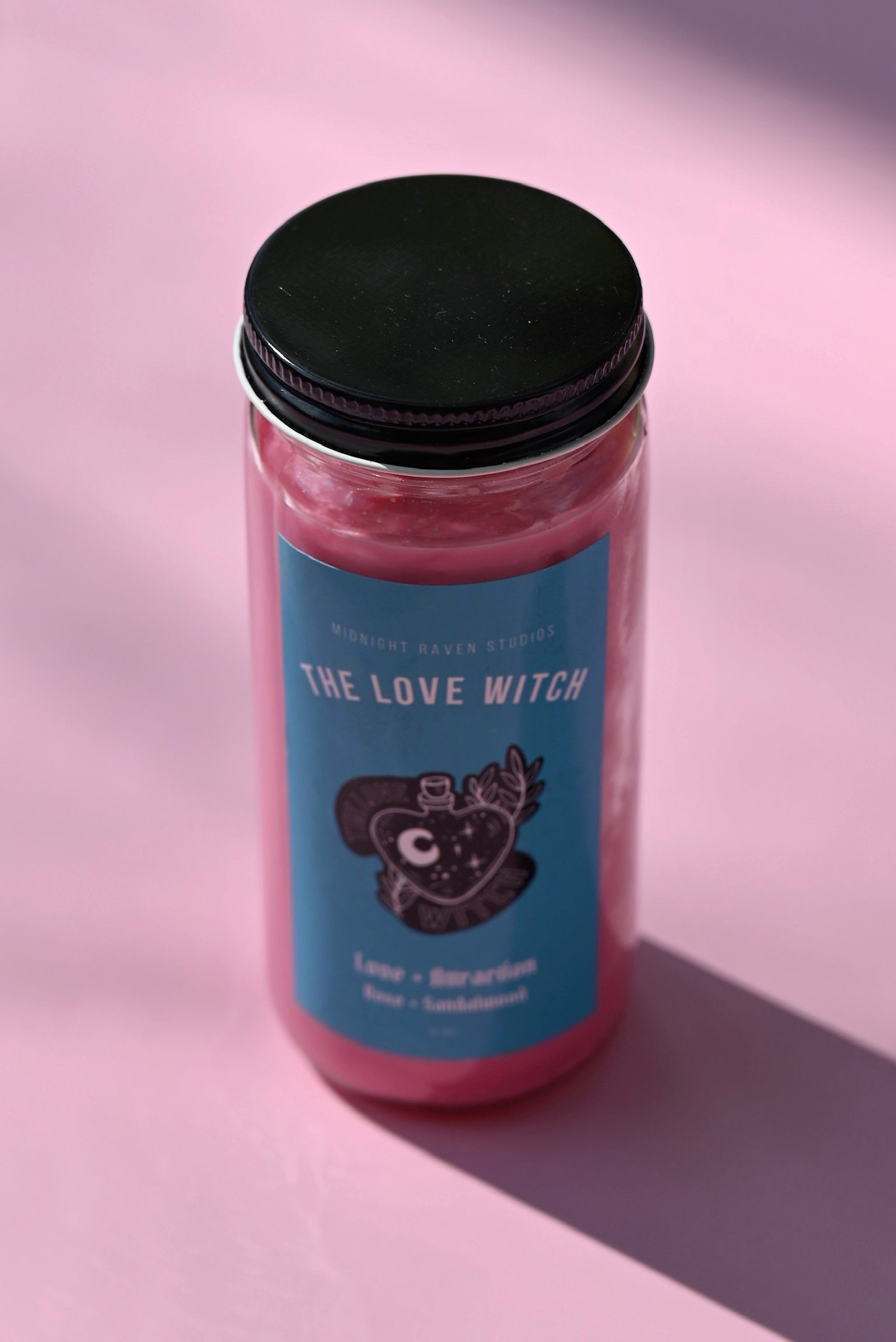 The Love Witch Spell Candle with Herbs and Crystals on a pink background.