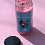 The Love Witch Spell Candle with Herbs and Crystals on a pink background with its lid to the side.