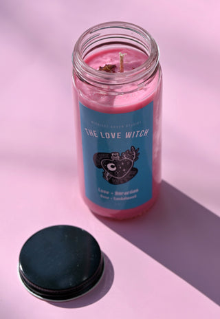 The Love Witch Spell Candle with Herbs and Crystals on a pink background with its lid to the side.